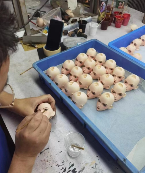 Artist hand-painting a tray of PU doll heads in the LORIBEAR workshop